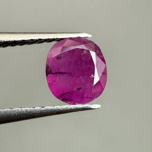 Könnte beinhalten: Ein facettierter, ovaler Rubin-Edelstein, gehalten von einer Pinzette. Der Rubin zeigt eine tiefe, gesättigte pink-rote Farbe mit inneren Einschlüssen. Der Edelstein ist poliert und reflektiert das Licht, wodurch seine Facetten zur Geltung kommen.