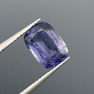 Könnte beinhalten: Ein rechteckiger, facettierter Edelstein, der von einer silbernen Pinzette gehalten wird. Der Edelstein hat eine tiefblaue bis violette Farbe mit helleren, durchscheinenden Bereichen. Die Facetten fangen das Licht ein und erzeugen einen funkelnden Effekt. Der Hintergrund ist einfarbig grau.