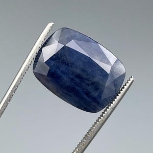 May include: A faceted, deep blue gemstone held by silver tweezers. The gem has a rectangular cushion cut with multiple reflective surfaces. The color is a rich, dark blue, with a slight variation in tone across the facets.