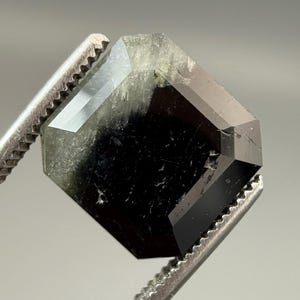 May include: A faceted, octagonal gemstone with a dark, almost black center and translucent, light green edges. The gem is held by a pair of silver tweezers, showcasing its geometric shape and polished surfaces.