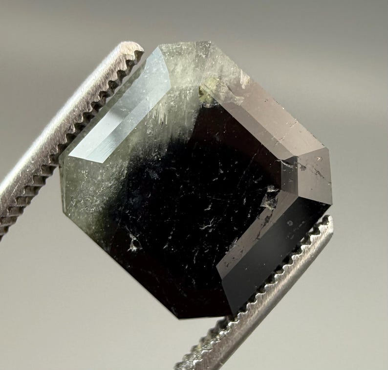 May include: A faceted, octagonal gemstone with a dark, almost black center and translucent, light green edges. The gem is held by a pair of silver tweezers, showcasing its geometric shape and polished surfaces.