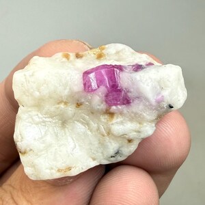 Könnte beinhalten: Ein rohes, natürliches Mineralexemplar mit einem leuchtenden, fuchsiafarbenen Kristall, eingebettet in eine cremeweiße Matrix. Der Stein hat ein rohes, unpoliertes Aussehen mit kleinen goldenen und schwarzen Flecken.