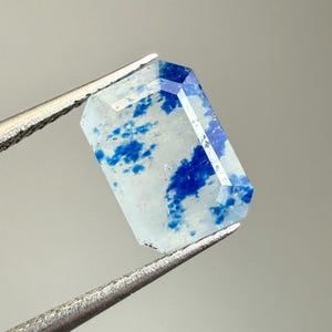 Könnte beinhalten: Ein rechteckiger Edelstein mit einer transparenten, hellblauen Basis und lebendigen blauen Einschlüssen. Der Stein wird von einer silbernen Pinzette gehalten, die seine facettierten Kanten und polierte Oberfläche zeigt. Das Design ist eine Mischung aus klaren und blauen Farbtönen.