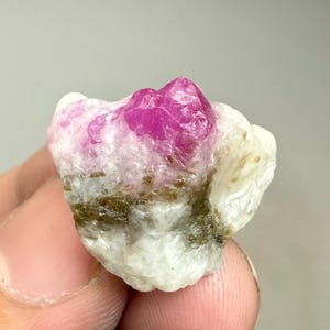 Könnte beinhalten: Ein rohes, natürliches Rubin-Kristall-Exemplar. Der Rubin hat eine leuchtende rosa-rote Farbe mit einer kristallinen Struktur. Er sitzt auf einer Matrix aus weißen und beigen Mineralien mit einigen grünen und braunen Einschlüssen.