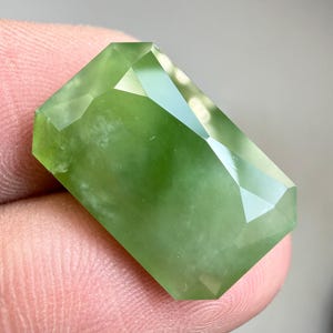 May include: A light green, rectangular gemstone with multiple facets. The cut of the gem creates a sparkling effect as it reflects light. The stone is a long rectangle with angled edges, showcasing its clarity.