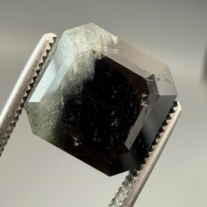 May include: A faceted, dark gemstone with a mix of black and green hues. The gem is held by a pair of metal tweezers, showcasing its geometric shape and polished surfaces. The gem's facets reflect light, highlighting its clarity and depth.
