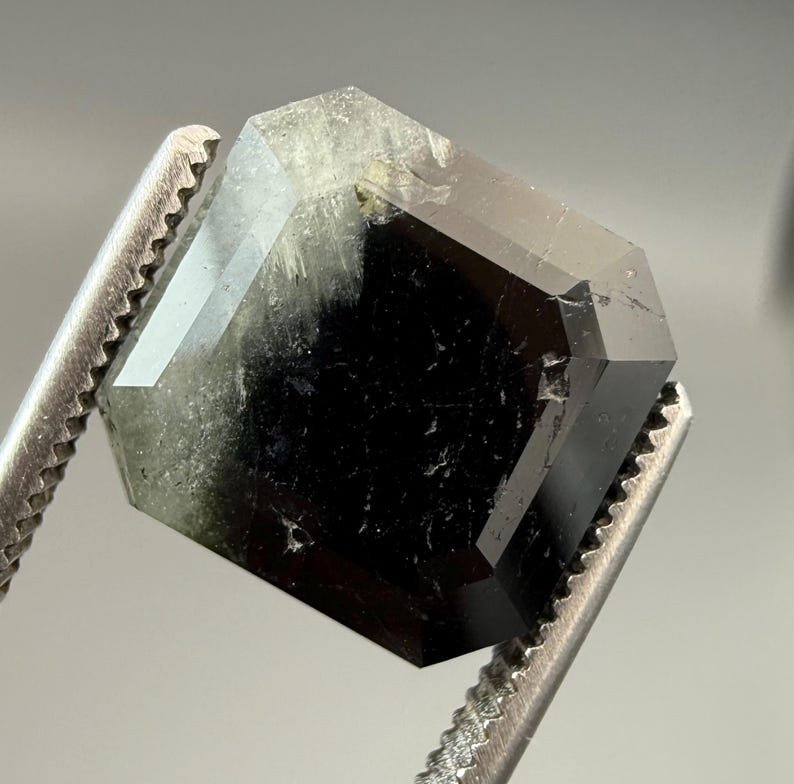May include: A faceted, dark gemstone with a mix of black and green hues. The gem is held by a pair of metal tweezers, showcasing its geometric shape and polished surfaces. The gem's facets reflect light, highlighting its clarity and depth.