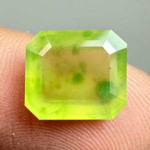 Peut inclure: Une pierre précieuse rectangulaire à facettes d'un vert citron vif et translucide. La gemme présente des inclusions internes et une surface polie, mettant en valeur sa clarté et sa brillance. La pierre est tenue par un doigt.