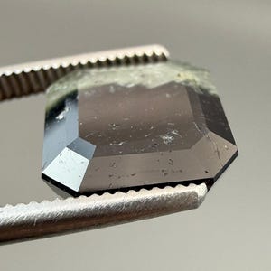 May include: A faceted, dark gray gemstone held by metal tweezers. The gemstone has an emerald cut with a slightly rough, green-tinged top edge. The facets reflect light, creating a sparkling effect. The tweezers have a serrated edge.