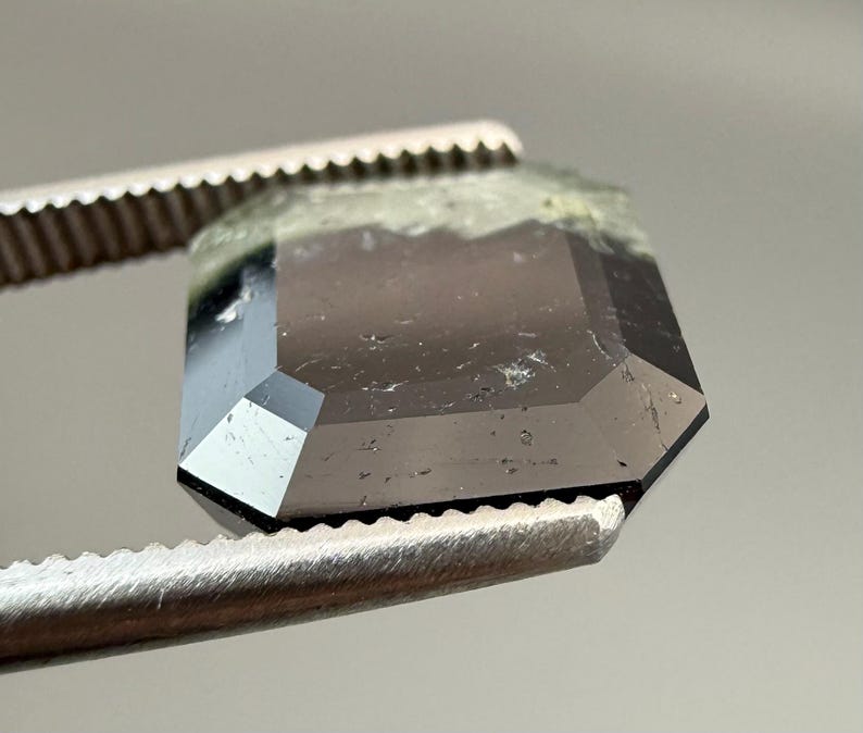 May include: A faceted, dark gray gemstone held by metal tweezers. The gemstone has an emerald cut with a slightly rough, green-tinged top edge. The facets reflect light, creating a sparkling effect. The tweezers have a serrated edge.