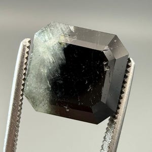 May include: A faceted, emerald-cut gemstone held by a pair of silver tweezers. The gem is a deep, dark green color with some lighter, translucent areas. The facets create a geometric pattern.