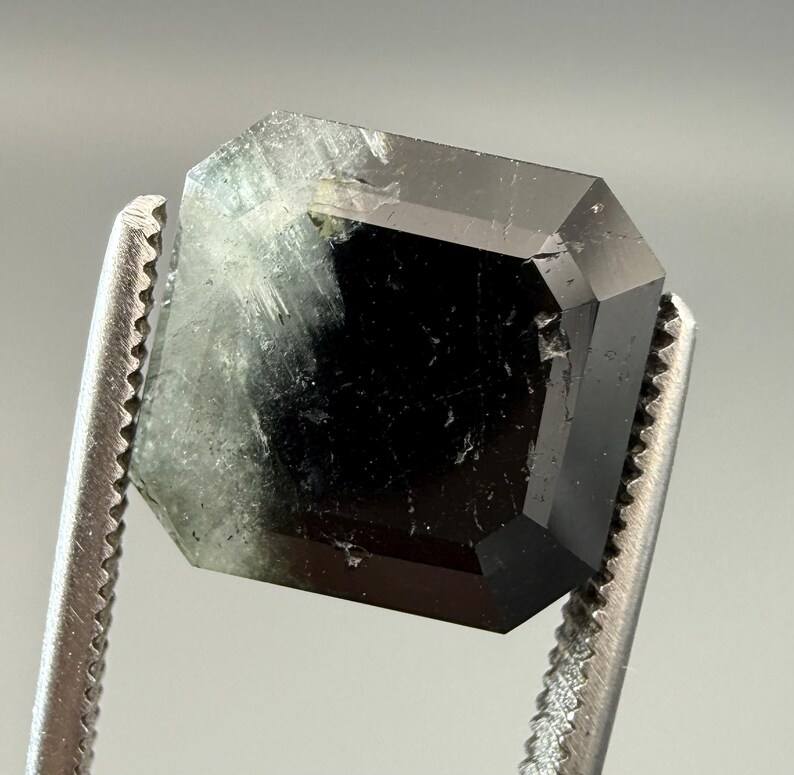 May include: A faceted, emerald-cut gemstone held by a pair of silver tweezers. The gem is a deep, dark green color with some lighter, translucent areas. The facets create a geometric pattern.