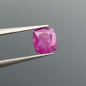 Puede incluir: Una vibrante gema rosa, talla cojín, sujeta por pinzas plateadas sobre un fondo gris. La gema tiene una superficie facetada, que capta la luz y muestra una gama de tonos rosados. Las pinzas dan una idea de la escala y el contexto.