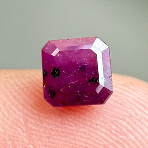 May include: A faceted, rectangular ruby gemstone with a deep magenta hue. The gem has a polished surface with small black inclusions. The cut is an emerald cut, with a flat top and beveled edges.