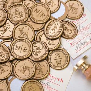 Puede incluir: Una colección de sellos de cera dorados con varios diseños de monogramas, incluyendo iniciales como "N/R" y "K&A". Algunos sellos son redondos, otros ovalados u ornamentados. La imagen también muestra tarjetas de eventos y un sello de cera.