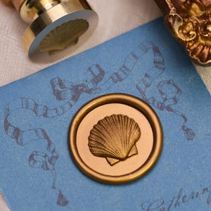 Puede incluir: Un sello de cera dorado con forma de concha marina sobre una tarjeta azul claro con un diseño de cinta. Se ve un sello de concha marina a juego. El sello de cera está presionado sobre la tarjeta, creando una impresión detallada. El tema es papelería y artesanía.