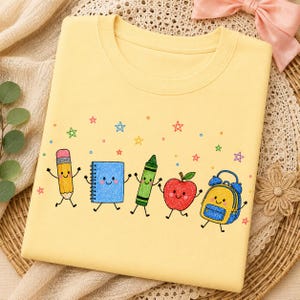 Könnte beinhalten: Ein hellgelbes T-Shirt mit einem bunten Grafikdruck von fünf anthropomorphen Schulsachen, darunter ein Bleistift, ein blaues Notizbuch, ein grüner Wachsmalstift, ein roter Apfel und ein blau-gelber Rucksack, umgeben von kleinen Sternen.
