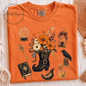 Pode incluir: Uma t-shirt laranja com uma estampa gráfica de uma bota preta cheia de flores laranjas e brancas, rodeada por uma bola de cristal, uma chave dourada, um livro preto, uma lanterna com vela, um corvo preto e um frasco de vidro com poção.