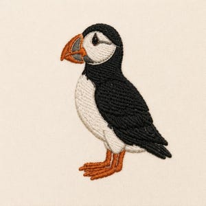 May include: An embroidered patch of an Atlantic puffin featuring a black back and wings, a white chest and face, a distinctive orange and grey beak, and orange webbed feet. The embroidery uses dense, textured stitching on a cream fabric base.