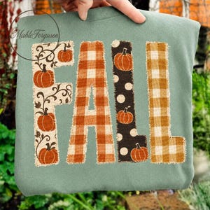 May include: A sage green sweatshirt featuring the word FALL in large, frayed-edge fabric letters. The F has a cream background with brown vines and orange pumpkins, the A is orange plaid, the L is black with white dots and pumpkins, and the L is gold plaid.