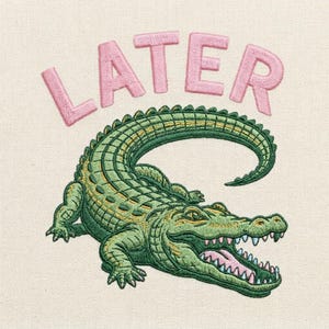 Puede incluir: Diseño bordado de un cocodrilo verde con la boca abierta y la palabra "LATER" en rosa encima. El cocodrilo tiene escamas detalladas y detalles amarillos. El diseño está sobre una tela color crema.