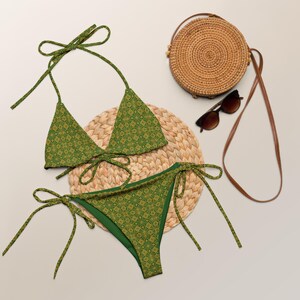 Könnte beinhalten: Grünes Bikini-Set mit geometrischem Muster, bestehend aus einem Triangel-Oberteil und einem Höschen zum Binden. Eine runde Korbtasche, Sonnenbrille und eine runde Flechtmatte ergänzen das Strand-Ensemble.