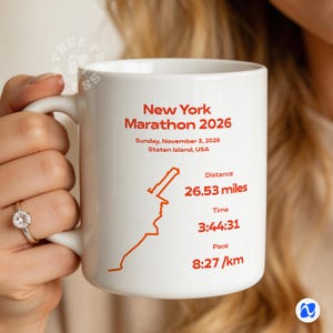 May include: White ceramic mug with the text "New York Marathon 2026" and race details: date, location, distance (42.69 km), time, and pace. The mug features a map outline of the race course.