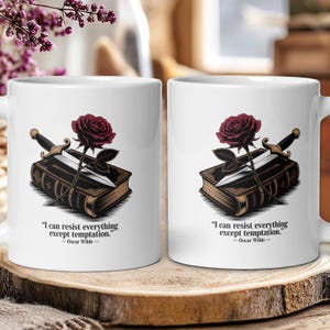 Puede incluir: Taza de cerámica blanca con un diseño de rosa roja, daga y libro. La taza tiene la cita "I can resist everything except temptation." de Oscar Wilde. La taza está sobre una superficie de madera.