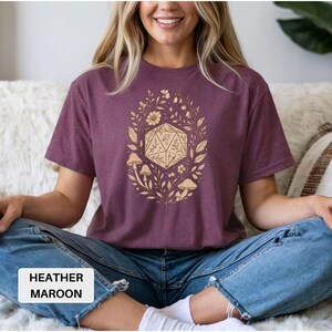 May include: Heather maroon t-shirt featuring a gold-colored design of a D20 die surrounded by leaves, flowers, and mushrooms. The text "HEATHER MAROON" is in a white rectangle at the bottom. The shirt is worn by a person.