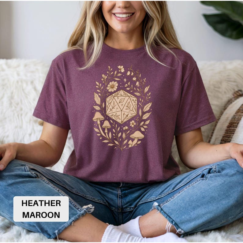 May include: Heather maroon t-shirt featuring a gold-colored design of a D20 die surrounded by leaves, flowers, and mushrooms. The text "HEATHER MAROON" is in a white rectangle at the bottom. The shirt is worn by a person.