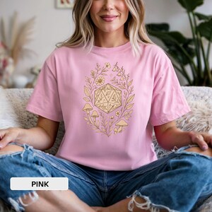 May include: Pink t-shirt featuring a beige D20 die surrounded by floral and mushroom illustrations. The text "PINK" is displayed at the bottom of the image. The shirt is made of a soft material.