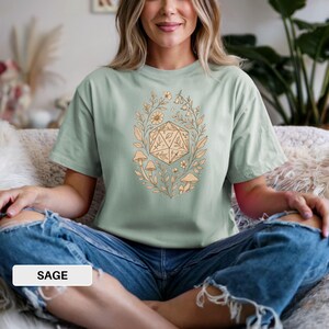 May include: Sage green t-shirt with a beige floral and geometric design. The design features a 20-sided die surrounded by flowers, leaves, and mushrooms. The word "SAGE" is printed on a white rectangle at the bottom of the image.