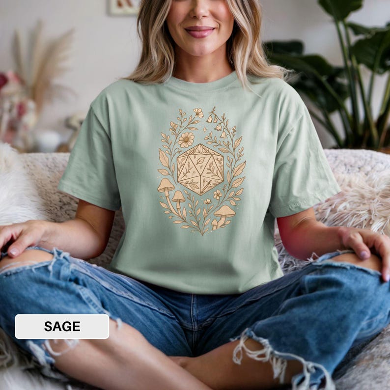 May include: Sage green t-shirt with a beige floral and geometric design. The design features a 20-sided die surrounded by flowers, leaves, and mushrooms. The word "SAGE" is printed on a white rectangle at the bottom of the image.