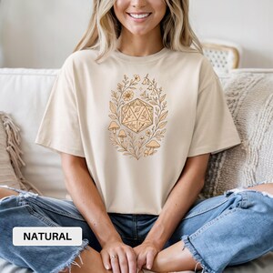 May include: Beige t-shirt with a nature-themed graphic. The design features a geometric shape surrounded by leaves, flowers, and mushrooms. The word "NATURAL" is printed in a white rectangle at the bottom of the image.