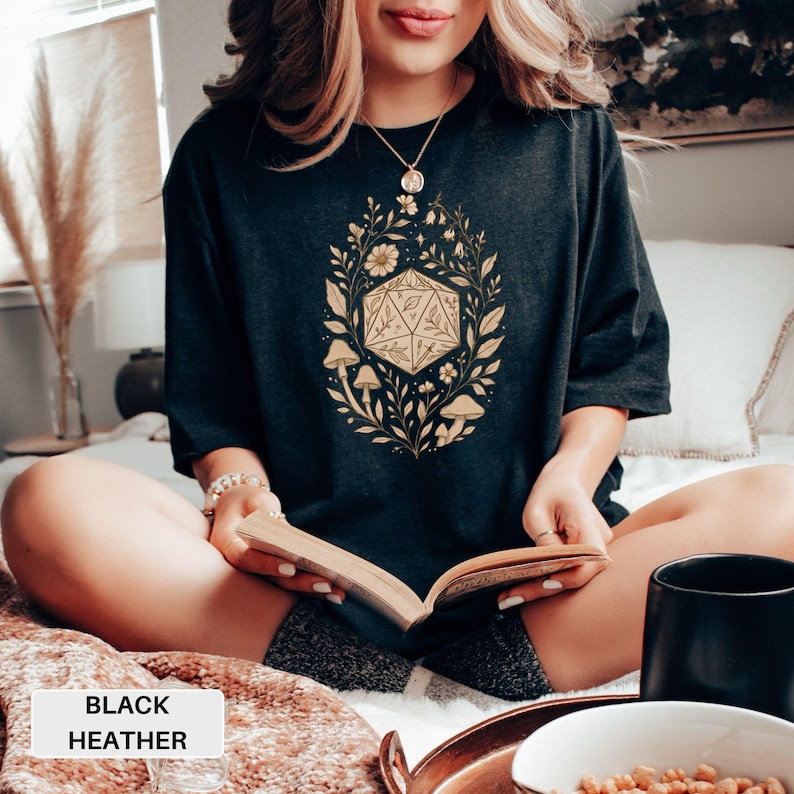 May include: Black heather t-shirt featuring a gold-colored D20 die surrounded by floral and mushroom illustrations. The text "BLACK HEATHER" is displayed on a white rectangle at the bottom of the image. The shirt is a dark gray color.