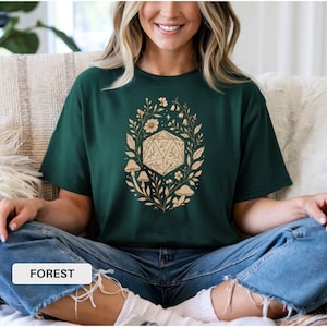 May include: Dark green t-shirt with a beige floral and geometric design. The design includes a D20 die, mushrooms, and foliage. The word "FOREST" is printed on a white rectangle at the bottom.