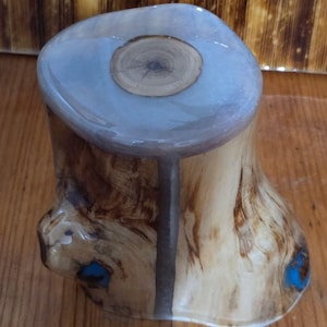 May include: A handcrafted wooden stool with a light blue resin top and a natural wood grain base. The base has a unique shape with a dark gray stripe and small blue accents. The top features a circular wood grain design.