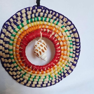May include: A circular woven wall decoration with a rainbow pattern. A small seashell hangs from the red central ring. The outer edge is a mix of purple, green, yellow, and natural tones. A black cord is attached for hanging.