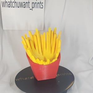 May include: A 3D-printed red container filled with yellow french fries. The container is shaped like a classic fast-food fry box. The text "whatchuwant_prints" is visible in the background.