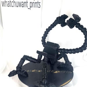 May include: Black 3D-printed phone holder with articulated arms and a circular base. The holder features the text "whatchuwant_prints" on a white background and on the base. The arms are flexible and can be adjusted to hold a phone.