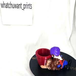 Op de afbeelding: Een grillig beeldje met een tuinkabouter met een paarse hoed en een blauwe broek, naast een rode bloempot. Het beeldje staat op een zwarte, ronde basis. De tekst "whatchuwant_prints" is zichtbaar.