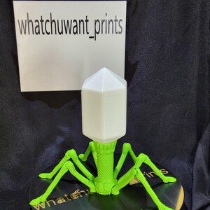 Pode incluir: Modelo impresso em 3D de um bacteriófago, com cabeça branca e pernas verdes. O modelo é exibido em uma superfície preta, com o texto "whatchuwant_prints" visível ao fundo.