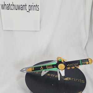 Könnte beinhalten: Ein Requisiten-Dolch mit schwarzem Griff, goldenen Akzenten und einer silbernen Klinge. Der Dolch ruht auf einer schwarzen, kreisförmigen Basis. Der Text "whatchuwant_prints" ist auf einer weißen Karte und auf der Basis zu sehen.