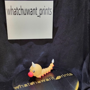 May include: A small, yellow and orange figurine of a cartoonish caterpillar with a red nose and a white horn. The figurine is on a black surface with the text "whatchuwant_prints". A white sign with the text "whatchuwant_prints" is in the background.