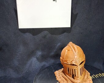 May include: A bronze-colored, 3D-printed knight's helmet with intricate details, including a visor and decorative elements. The helmet sits on a black surface with the text "whatchuwant_prints". The background is dark.
