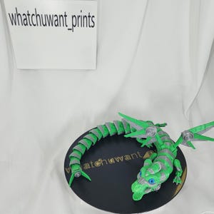 May include: A green and gray articulated dragon figurine with blue eyes, resting on a black circular base. The base has the text "watchuwant" printed on it. The dragon's wings and body are detailed.