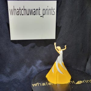 May include: A 3D-printed figurine of a woman in a yellow and white gown with her arms raised. The figurine is on a black surface with the text "whatchuwant_prints" on a white background and on the base.