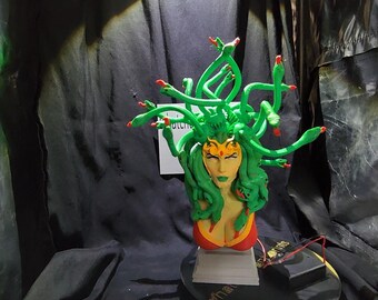 Puede incluir: Busto de Medusa verde y rojo, con serpientes verdes como pelo y un top rojo. La escultura está montada sobre una base negra con el texto "What Prints". El busto tiene una corona dorada.