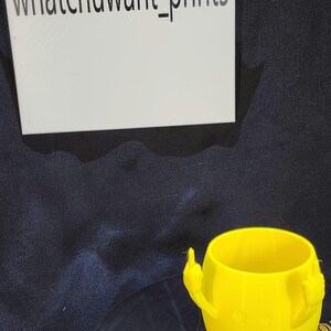 May include: A bright yellow 3D-printed cup with a smiley face and two middle fingers. The cup is sitting on a black surface. The text "whatchuwant_prints" is visible in the image.