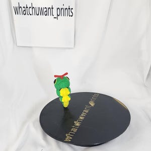 May include: A green and yellow caterpillar figurine with red antennae stands on a black, round display stand. The stand has the text "whatchuwant_prints" printed on it. The background is white, and the text "whatchuwant_prints" is also visible.